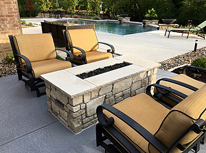 Concrete patio with black and beige furniture and rectangular gas fire pit with stone masonry and cut stone cap