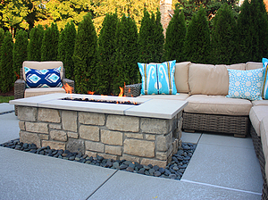 Concrete patio with beige furniture and rectangular gas burning fire pit with stone masonry and pewter mist cut stone cap