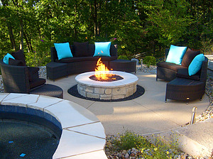 Concrete patio with black furniture and round gas fire pit with stone masonry and flagstone cap